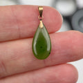 A teardrop-shaped green stone pendant with a gold bail, held between a person's fingers against a blurred background.